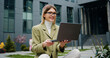 © serg - Outdoors portrait of smiling girl in glasses having conference call on laptop with sitting on street near business centre or university.