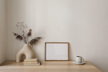 Modern organic shaped vase with dry flowers, grass, reed on old books. Cup of coffee, tea. Horizontal wooden picture frame, poster mockup on wooden table, desk. Home staging. Minimal interior.