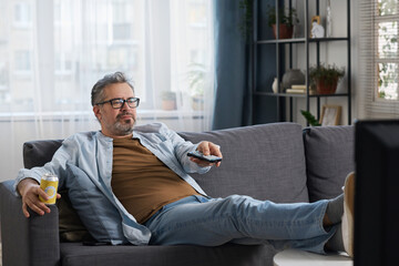 Wall Mural - Mature man with drink resting on sofa and switching channels with remote control while watching TV at home