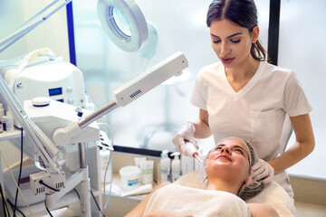 Wall Mural - Female beautician using cosmetology electrical too to apply darsonval facial massage to beautiful young woman at beauty clinic.
