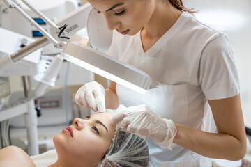 Wall Mural - Beautician squeezing acne and pimples from young woman's nose using lamp and magnifying glass in beauty spa clinic.