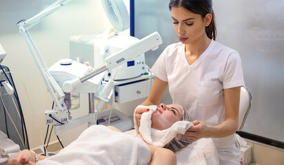 Wall Mural - Beautician removing clay mask from female client face with white towel during spa massage treatment at beauty clinic.