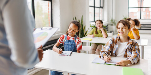 Wall Mural - Classmates sit at desks and listen to teacher during school lesson