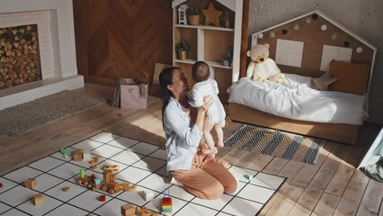 full shot of happy mother sitting on floor in cozy nursery, holding on arms and lifting up cute baby