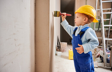 Little kid construction worker painting wall with brush in apartment under renovation. Child wearing safety helmet and work overalls while working on home renovation.