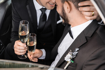 Smiling homosexual groom in formal wear holding champagne glass and hugging bearded boyfriend while celebrating wedding in car during honeymoon