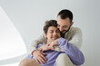 © LIGHTFIELD STUDIOS - Young brunette gay man hugging carefree boyfriend with closed eyes, in braces and in purple sweatshirt while sitting together on grey background with sunlight