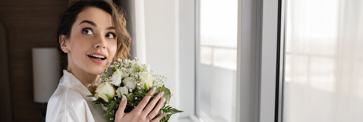 amazed woman with engagement ring on finger standing in white silk robe and holding bridal bouquet while looking up next to window in hotel suite, special occasion, bride on wedding day, banner
