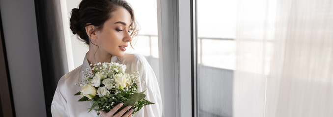 young woman with brunette hair standing in white silk robe and holding bridal bouquet next to tulle curtain and window in hotel suite, special occasion, bride on wedding day, banner