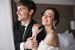 joyful bride in elegant jewelry and wedding dress hugging shoulder of happy groom in classic formal wear while standing together in modern hotel room after ceremony