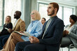 © pressmaster - Young serious businessman in formalwear using tablet while sitting among other learners at seminar or training and listening to lecture of coach