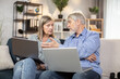 © sofiko14 - Gray-haired male in mature age reaching out hand to computer held by attractive senior lady sitting on couch in apartment. Married couple in jeans browsing online fashion store via internet app.