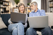 Gray-haired male in mature age reaching out hand to computer held by attractive senior lady sitting on couch in apartment. Married couple in jeans browsing online fashion store via internet app.