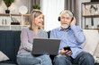 © sofiko14 - Cheerful senior pair having fun on comfortable sofa using modern laptop for watching comedy show in stylish living room. Smiling woman adjusting wireless earphones for beloved husband.