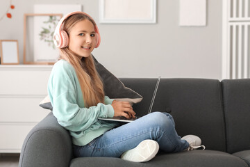 Wall Mural - Little girl in headphones using laptop on sofa at home