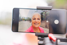 Through The Screen Of A Cell Phone An Older Latin Woman Vlogger Records A Video With A Gimbal Outside, Connecting With Her Audience On Social Networks