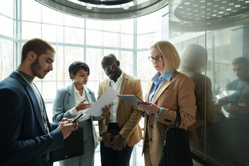 Poster - Group of serious colleagues in formalwear looking through their reports on papers and in gadgets before conference while moving in elevator
