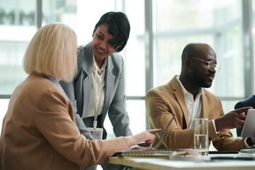 Poster - Young smiling businesswoman in formalwear looking at colleague pointing at laptop screen during discussion of online data at working meeting