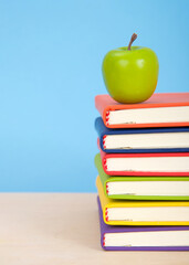 Bright colorful bound books stacked on a light wood table with blue background. Green apple on top of stacked books.