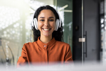 Portrait of Indian female programmer, woman in headphones listening to music, audio podcast, smiling and looking at camera, engineer developer closeup inside office at workplace, coding software.