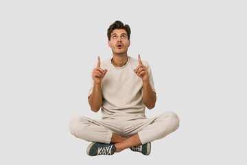 young caucasian man sitting on the floor isolated on white background pointing upside with opened mo