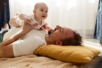 Happy dad holds his newborn baby in his arms while lying on the floor in a bright child's room. The father looks at the child with a loving look. Happy childhood and fatherhood.