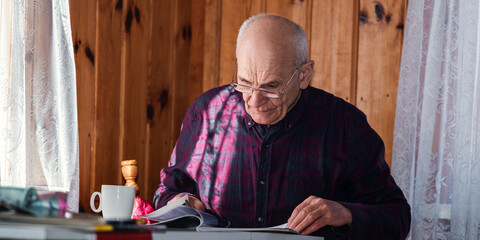 Older man reading newspaper seated at table inside of village house. Senior people eyesight problem. Old human leisure.