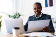 © Studio Marmellata - Concentrated Ethiopian male entrepreneur with laptop looking at document and analyzing data while sitting at desk and working on project in spacious workplace