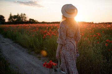 A girl in a white hat stands in a poppy field at sunset