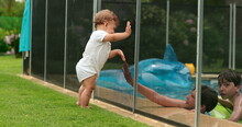 Baby Looking At Kids Play Inside Swimming Pool Water During Sumemr Day. Infant Leaning On Pool Fence