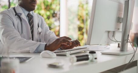 Wall Mural - Close Up of an African Male Doctor in White Coat Working on Computer at His Desk in the Office. Black Medical Health Care Professional Using Keyboard, Working with Patient Appointments