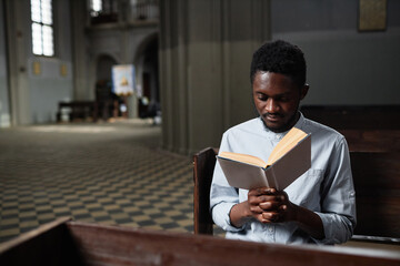 African American young man sitting on bench in church and reading prayer