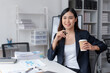 © amnaj - Successful Asian businesswoman smiling using laptop computer and holding coffee cup at office. Confident Asia businesswoman sitting happily in the office.