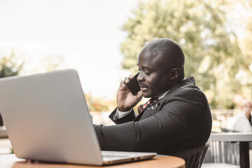 Sticker - Portrait of black african american businessman in suits sitting at a table in a summer cafe with a laptop and talking on the phone outdoors