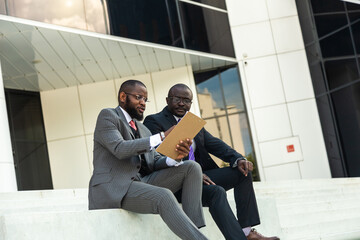 Sticker - Friendly meeting of business partners outdoors. Two dark-skinned men in suits sit on the steps of a city building with a notebook and have a conversation. Working break. Support and collaboration