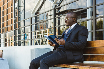 Sticker - A dark-skinned businessman in a suit reads an outdoor notebook while sitting on a bench against the backdrop of city buildings