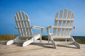 Sticker - Rear view of two white Adirondack style chairs sitting on a wooden deck, facing the beach. Vertical shot.