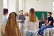 Smart and intelligent male teacher speaks in classroom surrounded by his high school students. Man talks to students at break or informal meeting where they discuss ideas about educational process.