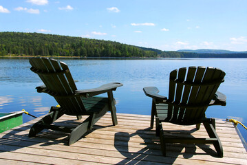 Poster - Two adirondack wooden chairs on dock facing a blue lake with clouds reflections