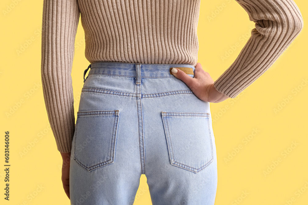 Young woman in stylish jeans on yellow background, back view