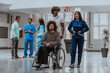 © Minet - In a modern hospital, a nurse and a technician compassionately transport a patient with disability while doctors conduct a discussion in a busy corridor, demonstrating teamwork and dedication in