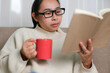 © Pornpimon - Happy young woman holding a cup of coffee and reading a book on the sofa after waking up in the morning at home. Lifestyle, free time and relaxation.