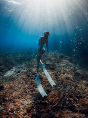 Wall Mural - Woman free diver glides underwater in clear sea. Freediving with beautiful girl in deep ocean and amazing sun rays