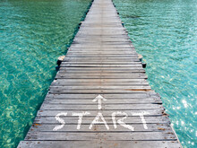 Start, White Text And Arrow On Wooden Bridge Heading To The Sea. Wood Plank Pathway Bridge On Clear Sea Water At The Port In Island On Sunny Day. Begin Weekend Holiday Vacation And Travel Background.