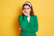 Bored sad offended woman wearing green jacket posing isolated over yellow background keeps hand under chin looking at camera with unhappy face displeased emotions.