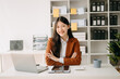 © Nuttapong punna - Young attractive Asian woman smiling thinking planning writing in notebook, tablet and laptop working from home, looking at camera at office