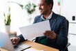 © Studio Marmellata - Cheerful elderly Ethiopian male executive manager with documents looking away and smiling while using laptop and working on corporate strategy in modern workspace