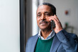 © Studio Marmellata - Positive aged Ethiopian male CEO director in formal suit speaking on mobile phone near window while working on successful project in spacious office