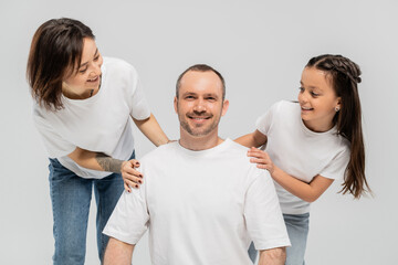 Wall Mural - woman with short hair and tattoo on hand and brunette teenage girl in blue denim jeans looking at man in white t-shirt on grey background, happy family