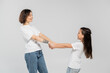 © LIGHTFIELD STUDIOS - happy mother with tattoo on hand and short hair holding hands with joyous daughter while standing together in white t-shirts and blue denim jeans on grey background, child protection day